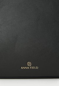 Black faux leather bag with a smooth texture, featuring a gold "ANNA FIELD" logo and a circular accent at the top.