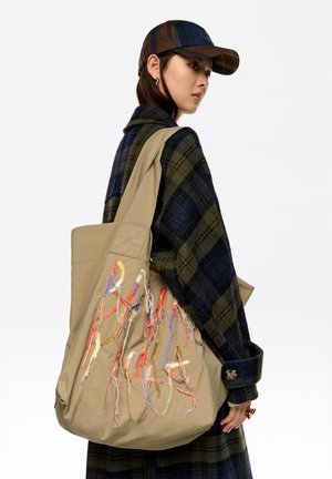 Bolso tote beige con bordado colorido, de gran tamaño, tela resistente y asas largas. Llevado sobre un abrigo de cuadros oscuros para crear contraste.