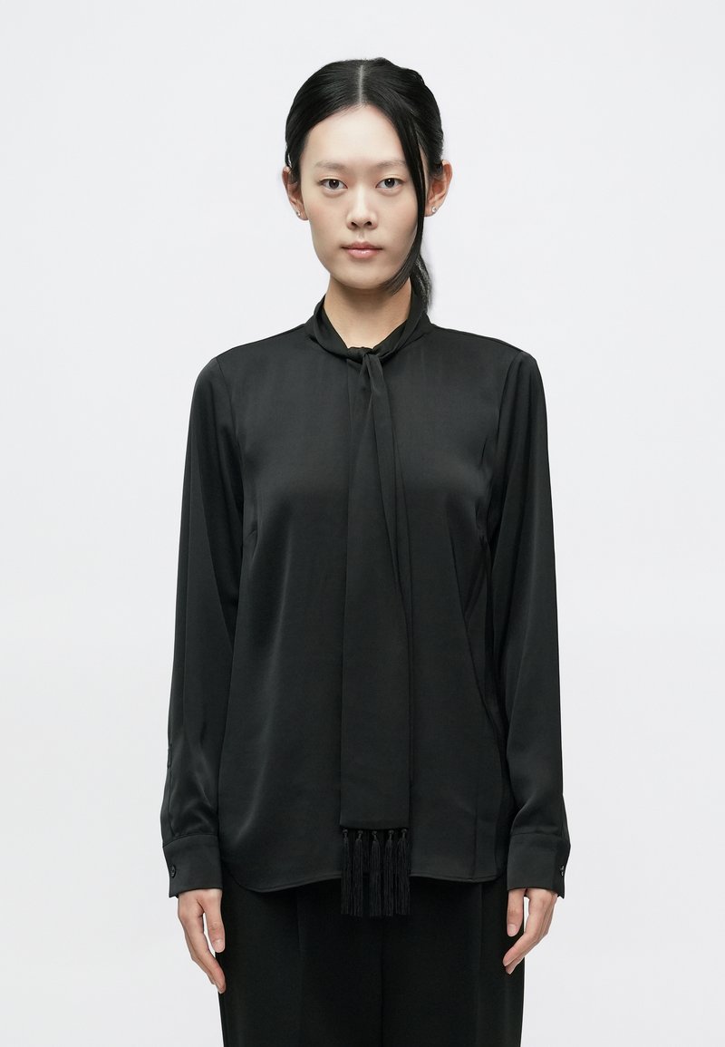 Čierna blúzka s dlhými rukávmi z hodvábneho materiálu s viazaním na krku a dekoratívnymi strapcami. Má gombíkové manžety a hladkú textúru po celom povrchu.