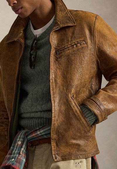 Καφέ δερμάτινο μπουφάν με vintage υφή, τσέπη στο στήθος με φερμουάρ και πλαϊνές τσέπες, φορεμένο πάνω από πράσινο πλεκτό πουλόβερ με σχέδιο πλεξούδας.