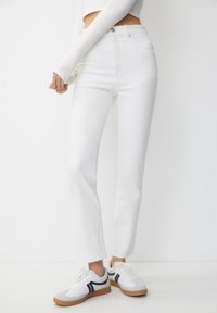 Žena v bílých džínech s vysokým pasem a bílých teniskách s černými pruhy, stojící na jednobarevném bílém pozadí.