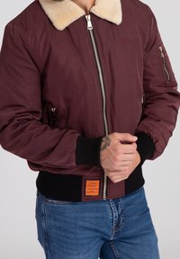 Mężczyzna w bordowej kurtce typu bomber z kołnierzem z baranka, czarnymi ściągaczami przy mankietach i u dołu oraz w niebieskich dżinsach, z rękami splecionymi na wysokości talii.