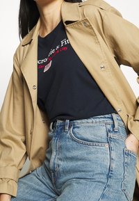 Chaqueta beige de botones sobre una camiseta navy con texto en blanco y rojo. Jeans de talle alto en azul claro con detalles en los bolsillos y textura desgastada.