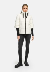 Witte pufferjack met hoge kraag, zwarte rits en trekkoorden. Heeft korte mouwen en padding, gecombineerd met zwarte skinny jeans en laarzen.