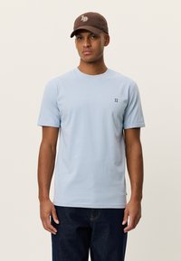 Ανοιχτό μπλε βαμβακερό μπλουζάκι με κοντά μανίκια, στρογγυλή λαιμόκοψη και μικρό σκούρο μπλε λογότυπο στο αριστερό στήθος. Φορεμένο με σκούρο τζιν και καφέ καπέλο.