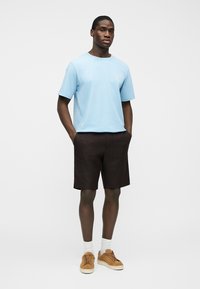 Homme debout les mains dans les poches, portant un t-shirt bleu clair, un short marron foncé, des chaussettes blanches et des baskets beige sur un fond uni.