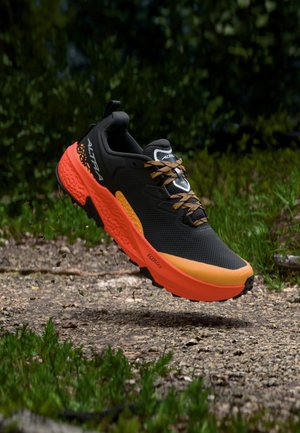 Zwart-oranje trailrunningschoen met robuust profiel zwevend boven een grindpad, omgeven door groene vegetatie.