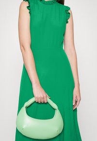Femme portant une robe verte sans manches tenant un sac à main vert clair à poignée tressée devant un fond uni.