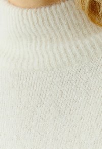 Pull en tricot couleur crème avec un col montant à côtes, présentant une texture douce et duveteuse ainsi que de subtils rayures verticales.