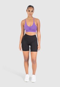 Violetti urheiluliivi, jossa on rypistetty etuosa, yhdistetty korkeavyötäröisiin mustiin shortseihin, molemmat sileästä, joustavasta materiaalista. Valkoiset lenkkarit.