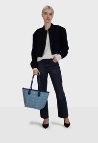 Niebieska torba typu tote z wzorem i granatowymi akcentami, trzymana przez osobę ubraną w czarną krótka kurtkę na białym swetrze i ciemne jeansy.