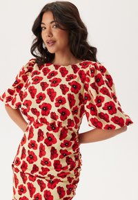 Robe florale à manches bouffantes, avec un fond crème et un motif de coquelicots rouges. Le tissu a une apparence texturée, accentuant le design.