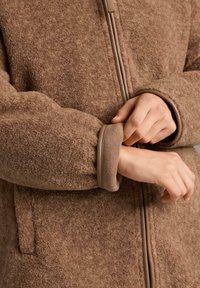 Hnedá fleecová bunda s jemnou textúrou, vybavená zapínaním na zips a rebrovanými manžetami. Doplnok tvorí hladký látkový lem pozdĺž zipsu.