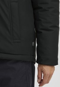 Chaqueta negra aislante con textura suave, bolsillo con cremallera y una etiqueta en el costado. Los puños están ligeramente acolchados y son redondeados.