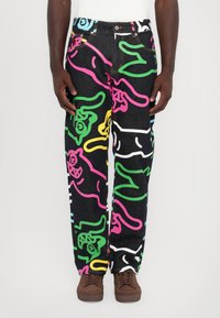 Denim pants featuring a black background with colorful, abstract cartoon-like patterns in pink, green, yellow, and blue. Brown shoes.