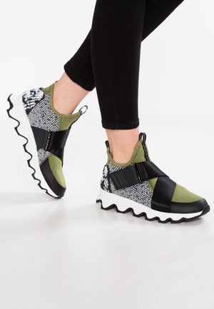 Personne portant des baskets à enfiler à motifs verts, noirs et blancs avec des semelles épaisses ondulées, et des leggings noirs, debout sur un sol clair.