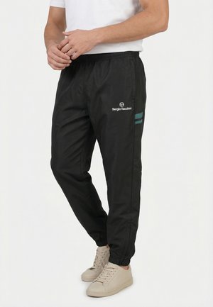 Man draagt zwarte Sergio Tacchini trainingsbroek met groene strepen en witte sneakers, staand tegen een eenvoudige lichte achtergrond.