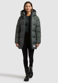 Puffervest i mørkegrå, med hætte, lynlås lukning og sidelommer. Båret over en grøn top og sorte leggings, med sorte støvler.
