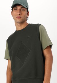 T-shirt in cotone verde scuro con maniche corte, caratterizzato da un design a diamante testurizzato e un piccolo logo sul petto.