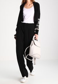 Cardigan nero leggero con dettagli a righe, abbinato a una canotta bianca e pantaloni neri larghi. Zaino grigio chiaro, sneakers bianche.