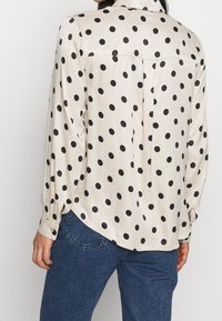 Blusa in satin con sfondo crema e grandi pois neri. Maniche lunghe, orlo curvo e dettaglio della spalla posteriore.