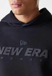 Felpa con cappuccio nera realizzata in tessuto morbido, caratterizzata da un prominente logo "NEW ERA" in rilievo e un piccolo logo sopra di esso. I bordi del cappuccio sono dettagliati e morbidi.