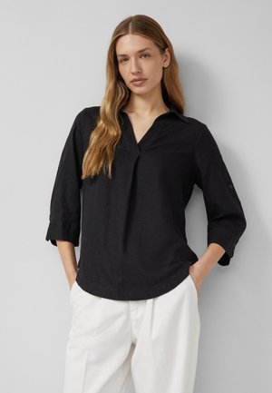 Femme aux longs cheveux blonds porte un chemisier noir à manches trois-quarts et un pantalon blanc, debout les mains dans les poches sur fond gris.