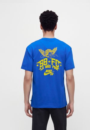 Person iført en blå Nike SB t-shirt med en gul engelgrafik og tekst "SB FC La Más Fina" på ryggen, stående mod en ensfarvet baggrund.