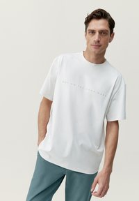 Biela bavlnená tričko s krátkymi rukávmi a okrúhlym výstrihom, s tlačou svetlošedej farby, na ktorej je nápis "HOP ON THE BORN WAVE."