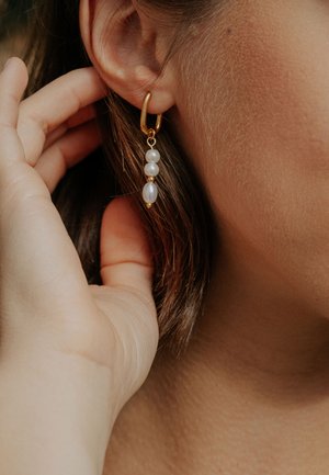 Prim-plan al unei mâini care ajustează o cercei cerc aurii cu trei perle albe pe urechea unei persoane cu păr castaniu.