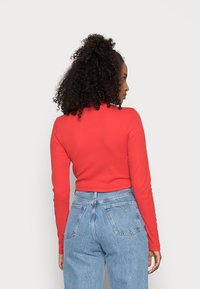 Blusa longa de mangas com nervuras vermelhas, cortada na cintura, combinada com jeans de ganga azul de cintura alta, com bolsos traseiros e um tom claro.