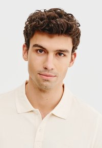 Beżowa koszulka polo z klasycznym kołnierzykiem, z zasuwaną plisą. Materiał wydaje się gładki z delikatną teksturą. Czysty, minimalistyczny design.