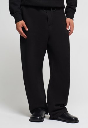 Persona che indossa pantaloni neri larghi con coulisse in vita e scarpe nere slip-on, in piedi davanti a uno sfondo semplice.