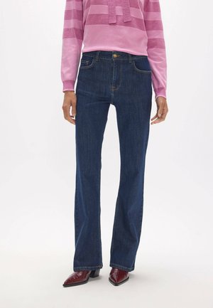 Persona que lleva jeans azul oscuro de pierna recta, un suéter rosa a rayas y botas de punta estrecha color rojo oscuro, de pie sobre un fondo blanco.