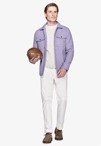 Lyse-lilla uldskjortejakke med to brystlommer over en hvid kabelstrikket sweater, parret med hvide bukser og brune ruskinds sko. Holder en brun fodbold.