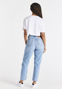 Eine Frau mit langen geflochtenen Haaren steht mit dem Rücken zur Kamera, trägt ein weißes, kurzes T-Shirt, helle Jeans und weiße Sneakers vor einem weißen Hintergrund.