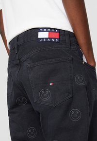 Black denim jeans with a TOMMY JEANS label, featuring a patterned design of circular NY logos. Includes back pockets and stitching details.