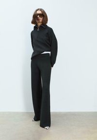 Crni pulover s polu-zip ovratnikom, uz široke crne hlače. Oba su izrađena od mekog materijala s glatkom teksturom.