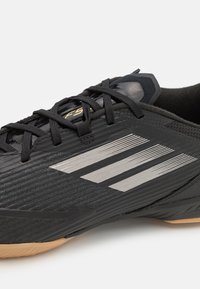 Zapatilla deportiva negra con parte superior texturizada, tres rayas plateadas y cordones negros; suela de goma negra y marrón, diseño elegante.