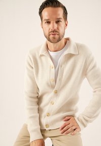 Cremefarbener gestrickter Cardigan mit Knopfleiste, geripptem Muster und spitzem Kragen, kombiniert mit einem weißen T-Shirt und hellen Hosen.