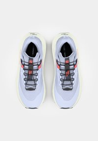 Baskets de sport gris clair avec une tige texturée, des lacets noirs et des accents orange vif. La semelle est rembourrée et légèrement surélevée.