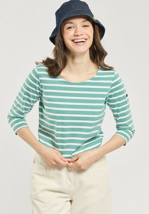 Jeune femme souriante, portant un chemisier rayé vert et blanc à manches trois quarts, un pantalon crème et un chapeau bob bleu foncé.