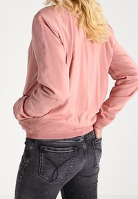 Jasnoróżowa bomberka z długimi rękawami, elastycznymi mankietami i pasem. Gładka tekstura, prosty fason i luźny krój. Noszona na ciemnych dżinsach.