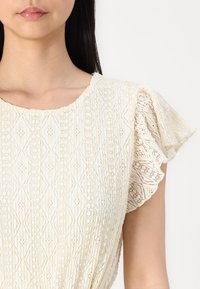 Blouse en dentelle de couleur crème doux avec un motif en diamant délicat, des manches à volants et un tissu texturé. Col rond et éclat subtil.