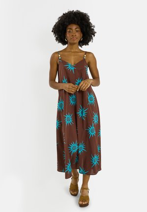 Mujer con cabello rizado natural que lleva un vestido maxi marrón sin mangas con patrones de sol azules y sandalias de plataforma marrones.