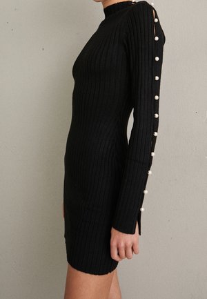 Kobieta ubrana w dopasowaną czarną dzianinową sukienkę z żebrowanym wzorem, ozdobioną perłowymi guzikami biegnącymi wzdłuż rękawa od ramienia do mankietu.