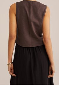 Kobieta ubrana w ciemnobrązowy bezrękawnik i czarną spódnicę, widziana od tyłu, z złotą bransoletą na lewym nadgarstku.