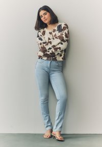 Džemper od pletene tkanine u krem nijansi s apstraktnim uzorkom u smeđoj i crnoj boji, uparen s pripijenim svijetloplavim trapericama i bež sandalama s trakama.