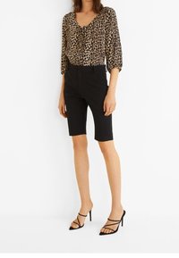Blouse imprimé léopard avec manches larges, associée à un short noir ajusté. Portée avec des talons noirs à lanières. Fond simple.