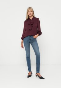 Blusa bordeaux con dettaglio a fiocco, maniche lunghe; abbinata a jeans attillati blu e scarpe con tacco nere. Sfondo semplice.